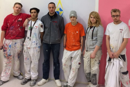Lehrlinge aus der Malerei gemeinsam mit Ausbildner vor Logo der Aids Hilfe © Wien Work